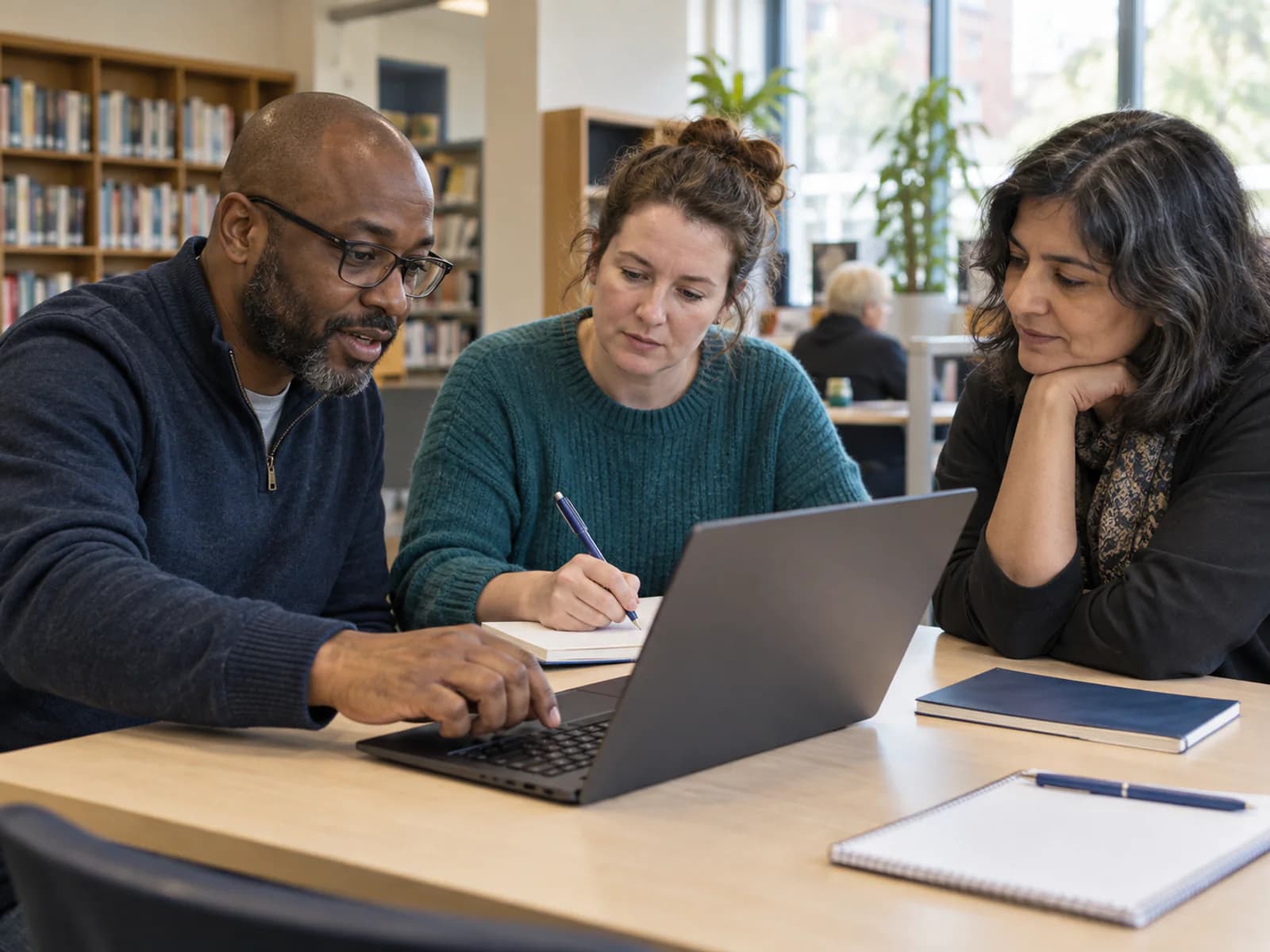 Three adults learning together with a laptop and notebooks in a library