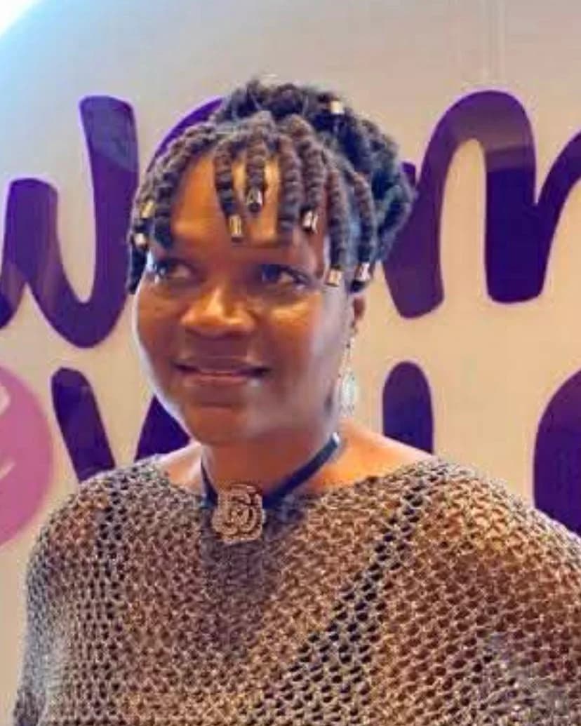 Laga Monique Deza smiling in front of a white wall with large purple lettering
